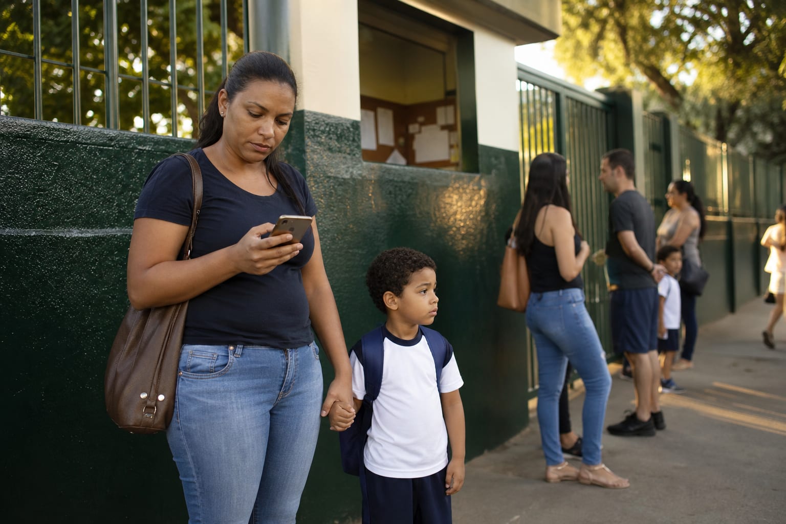 Mãe com filho uniformizado consulta o celular na calçada em frente à escola