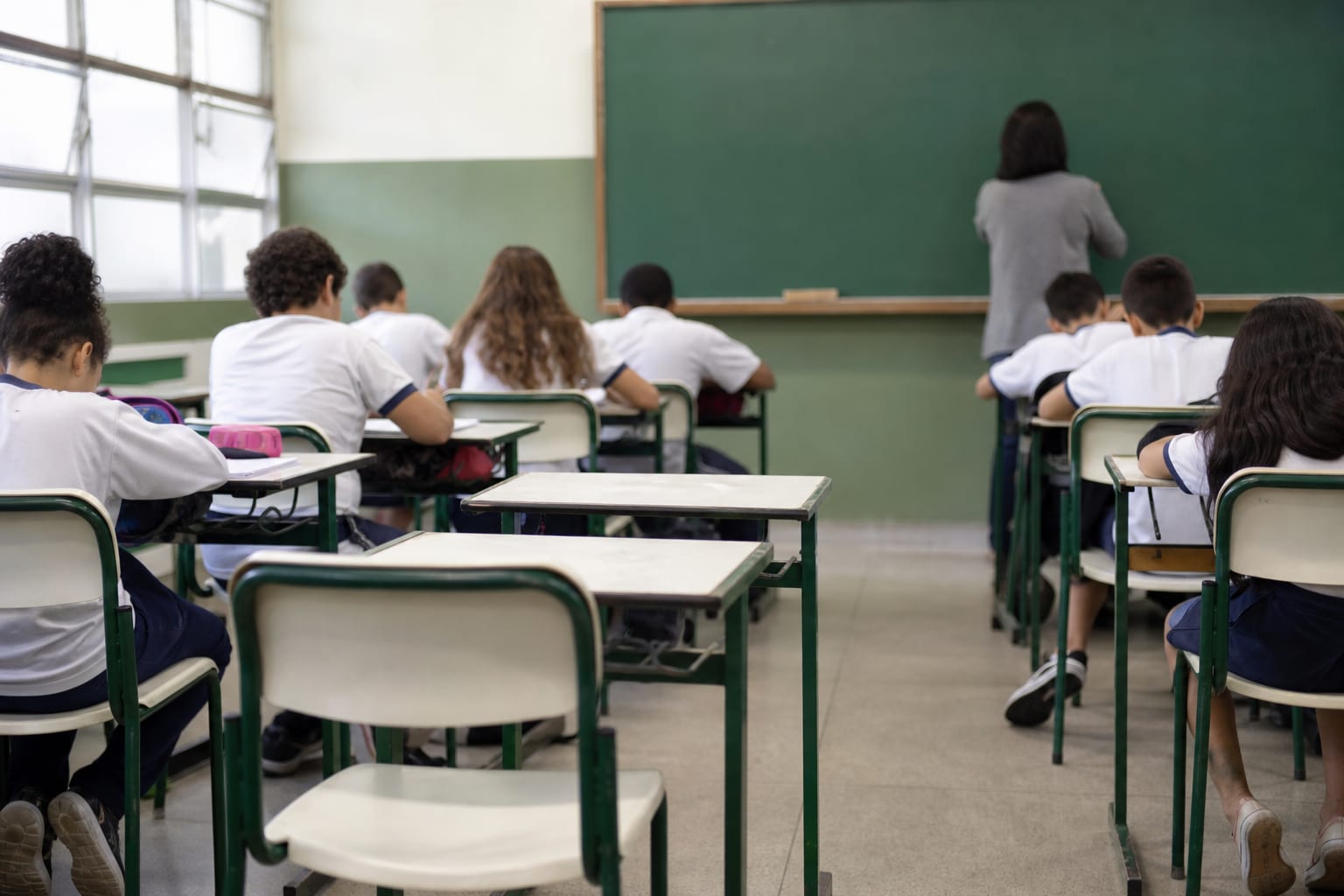 Carteiras vazias no meio de uma sala de aula com alunos uniformizados ao redor